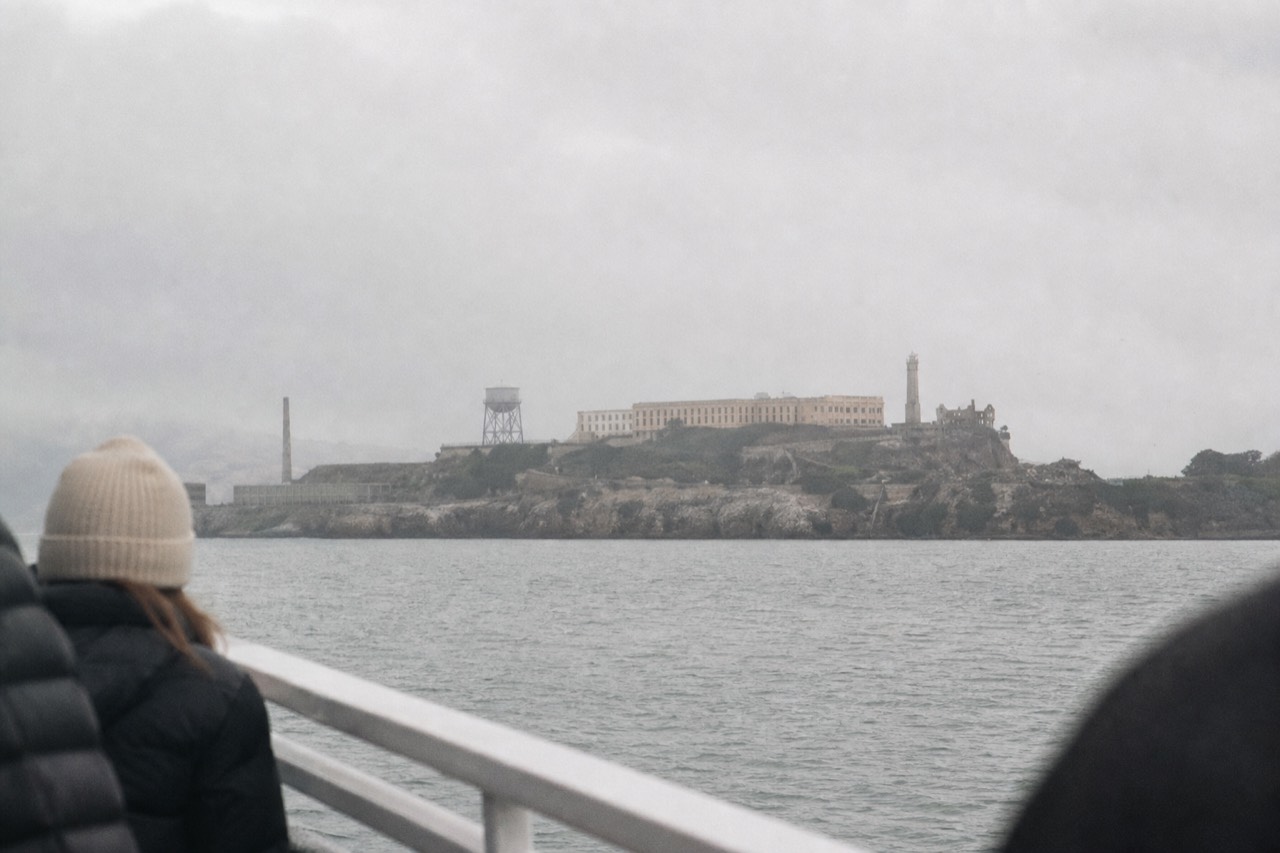 Passengers in silhouette along a boat rail at dusk facing Alcatraz, the island's lights beginning to glow across a darkening bay with the skyline faint behind.