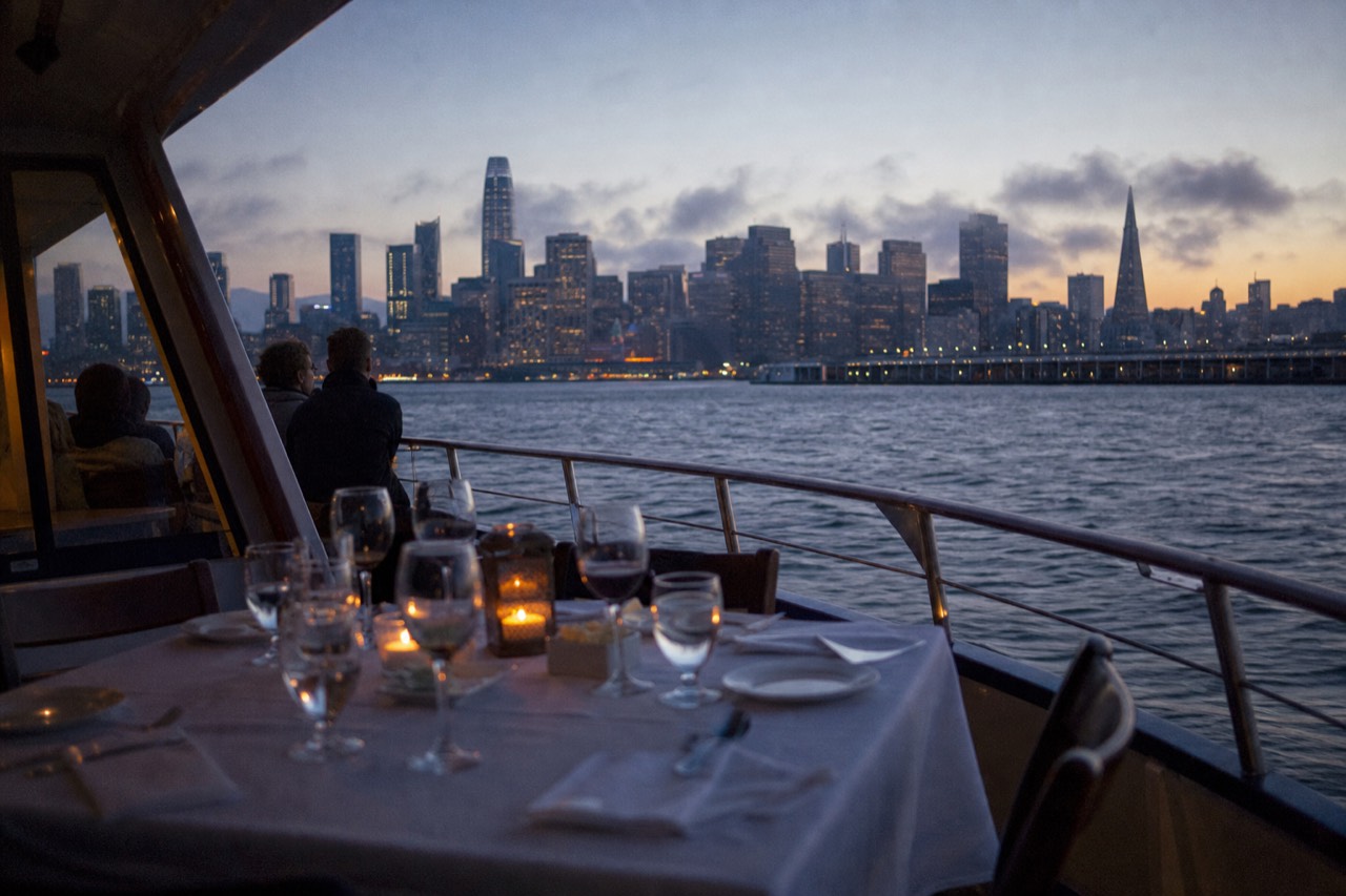 A warmly lit dining boat crossing dark bay water at nightfall, the downtown San Francisco skyline glowing behind it and reflections streaking the water.