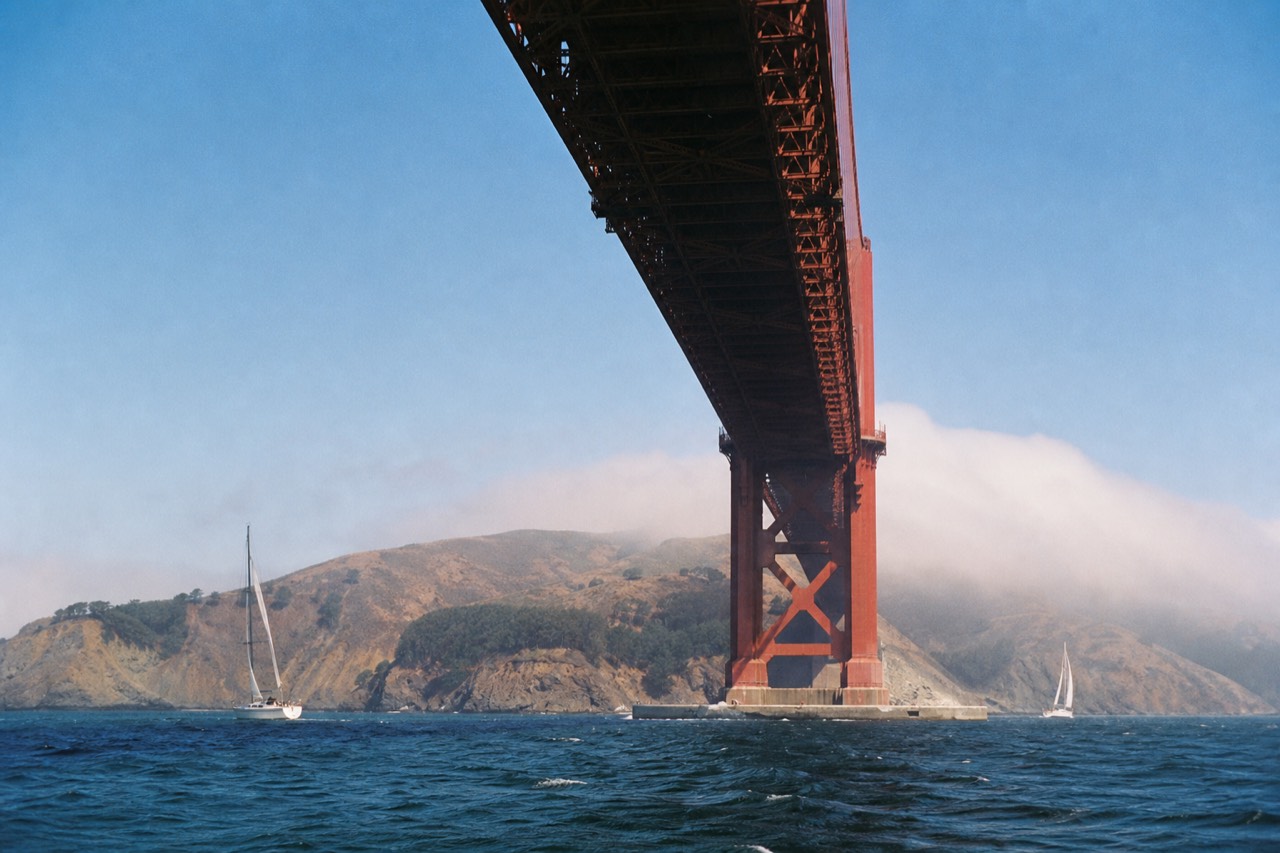 A sightseeing boat rounding below the north tower of the Golden Gate Bridge at golden hour, passengers along the rail, green headlands behind.