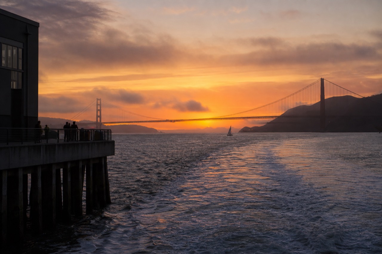 The downtown San Francisco skyline lit gold and pink at sunset seen low across calm bay water, a cruise boat's silhouette crossing the reflection.
