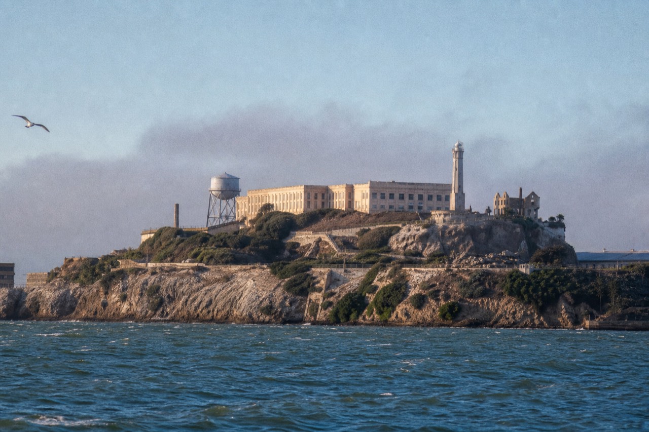 A white sightseeing boat rounding close beneath the steep east cliff of Alcatraz, the old cell-house windows and rusted railings visible above dark rock and breaking surf.