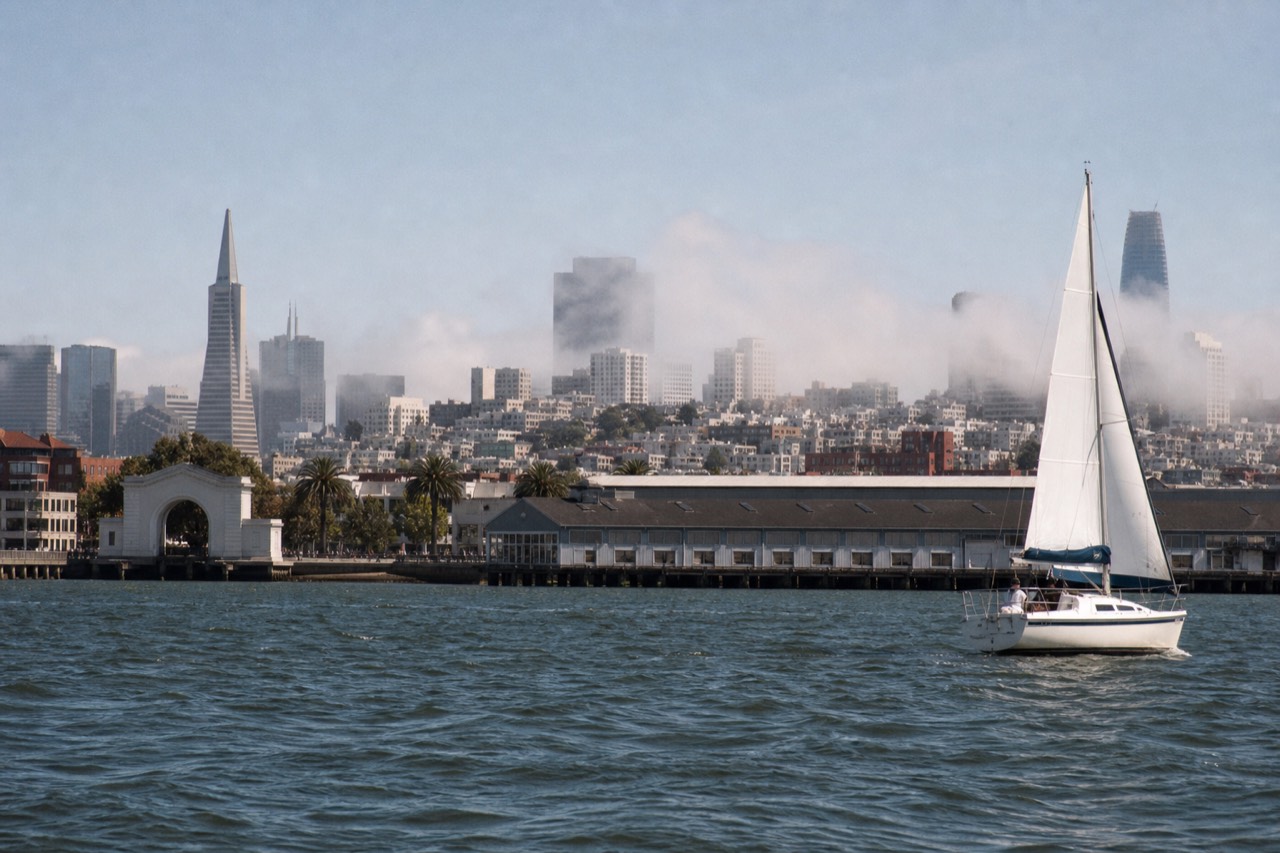 A white sailing catamaran heeling under full sail on a breezy bay, spray at the bow, a green island and distant bridge towers filling the background under bright sky.