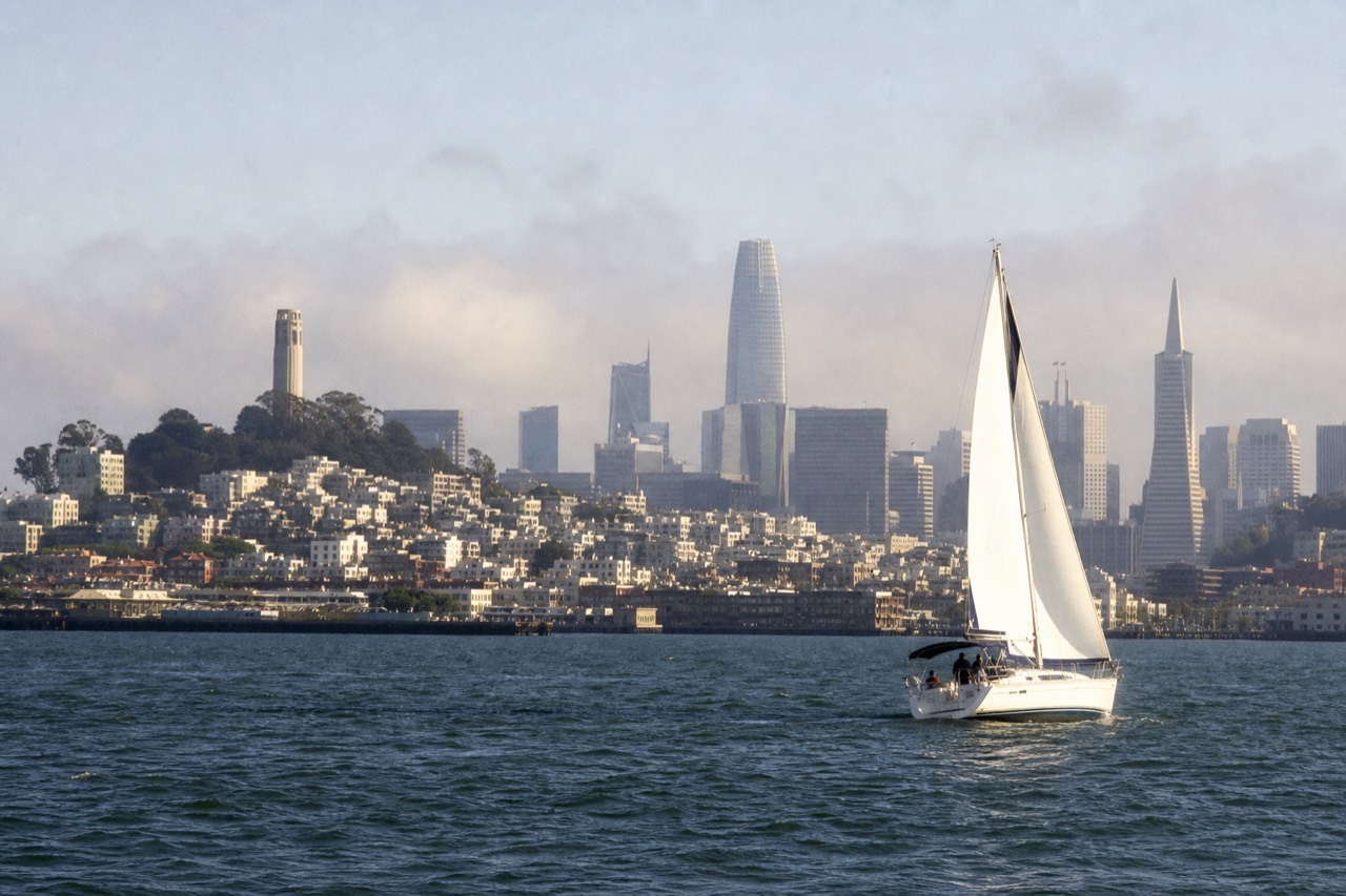Fog pouring low through the towers of the Golden Gate Bridge on a summer morning, a small sightseeing boat dwarfed on the pale grey water beneath.